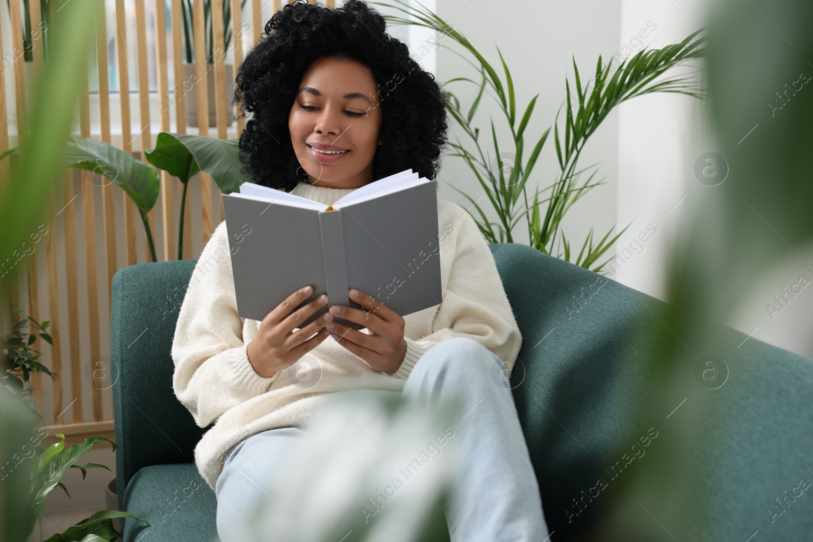 Photo of Relaxing atmosphere. Happy woman reading book on sofa surrounded by beautiful houseplants in room