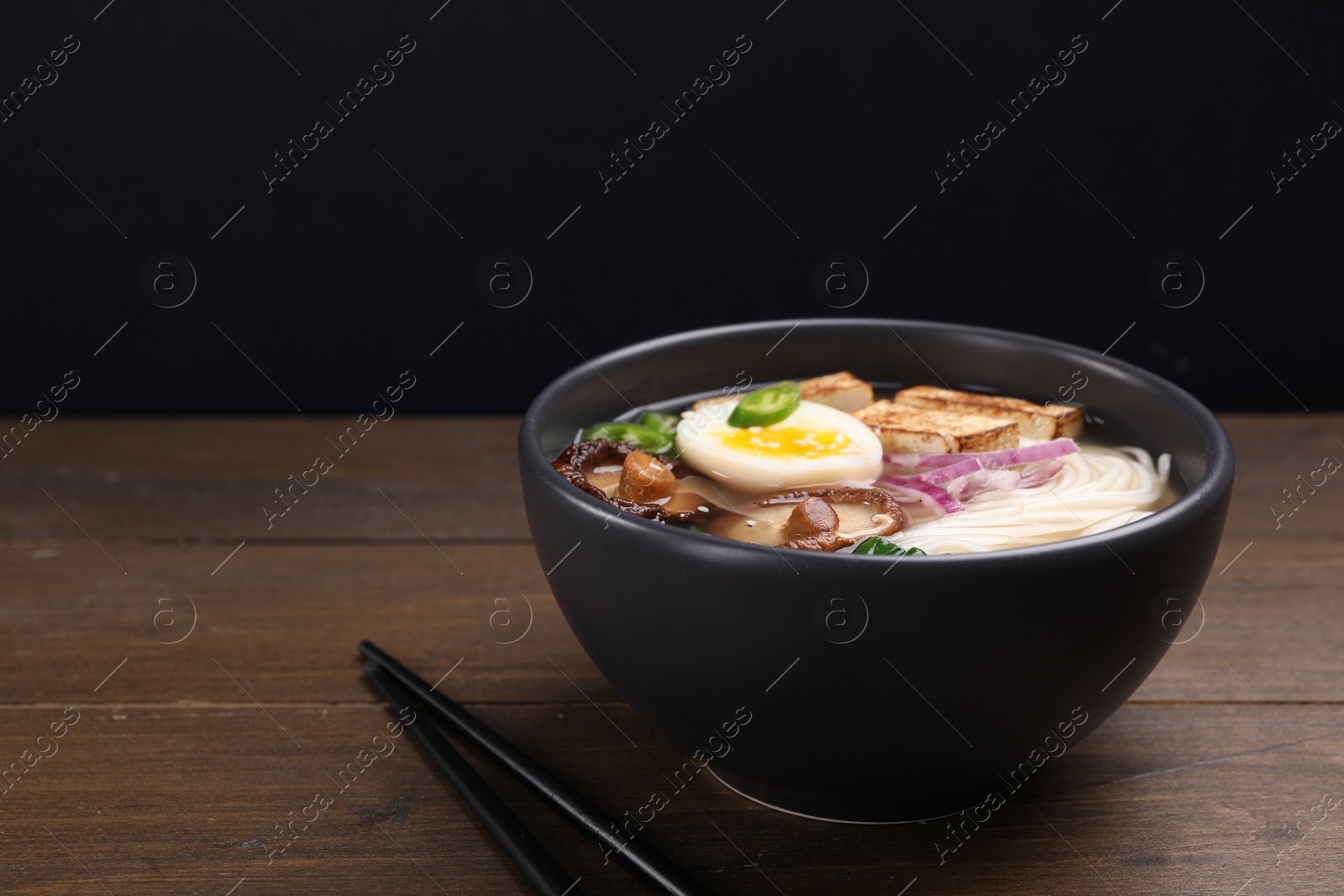 Photo of Delicious vegetarian ramen in bowl on wooden table against black background. Space for text