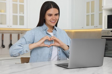 Happy young woman having video chat via laptop and making heart at table in kitchen