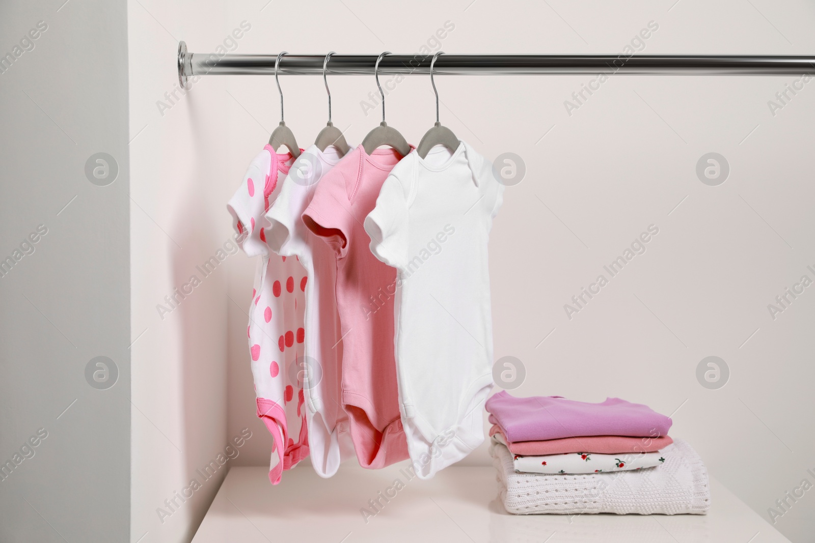 Photo of Hangers with baby bodysuits and stack of clothes near white wall