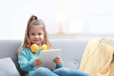 Photo of Cute little girl with headphones and tablet on sofa at home. Space for text