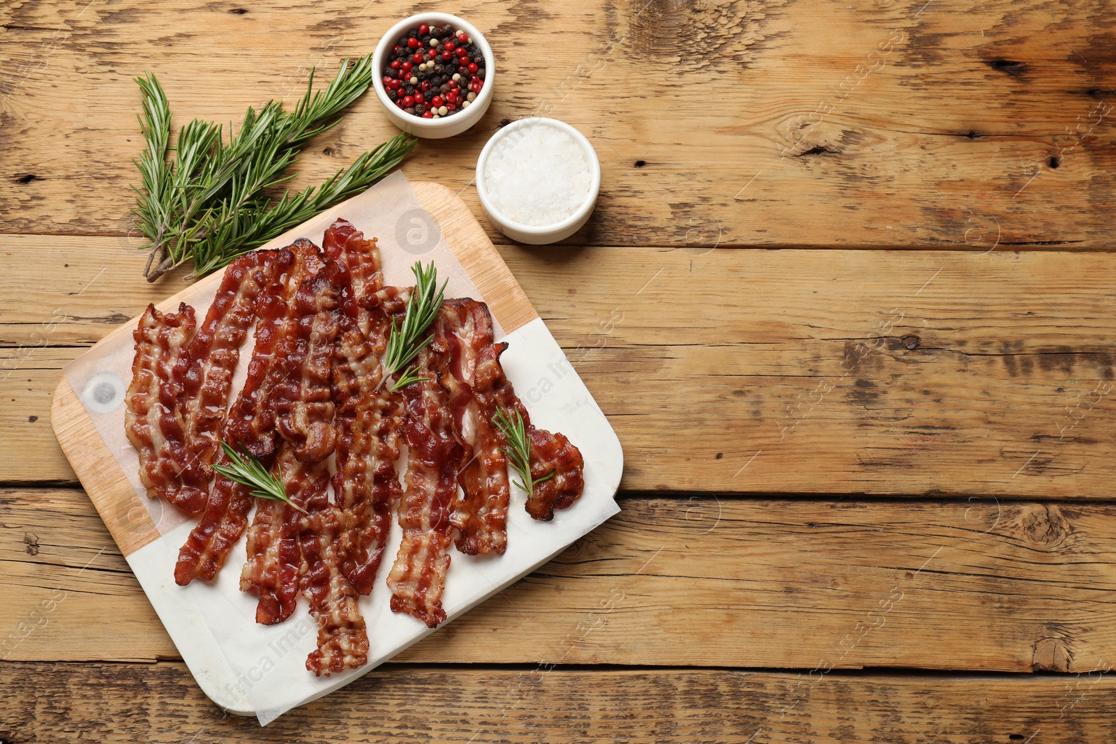 Photo of Slices of tasty fried bacon, rosemary, salt and peppercorns on wooden table, top view. Space for text