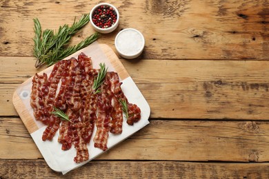 Photo of Slices of tasty fried bacon, rosemary, salt and peppercorns on wooden table, top view. Space for text