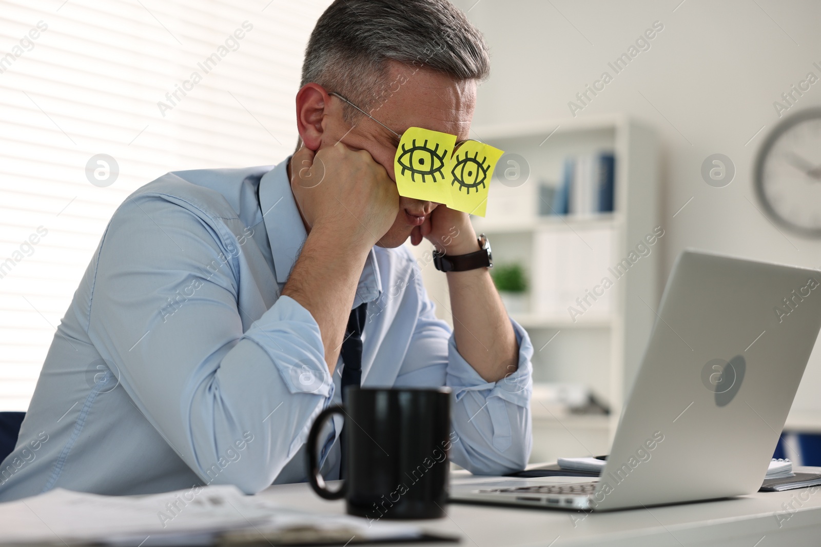 Photo of Man with fake eyes painted on sticky notes snoozing at workplace in office