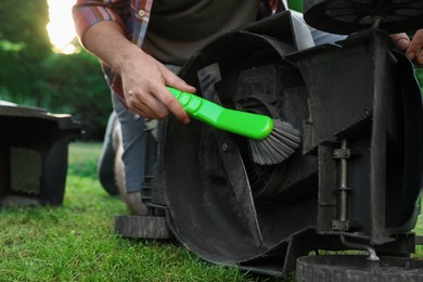 Photo of Man cleaning lawn mower with brush in garden, closeup