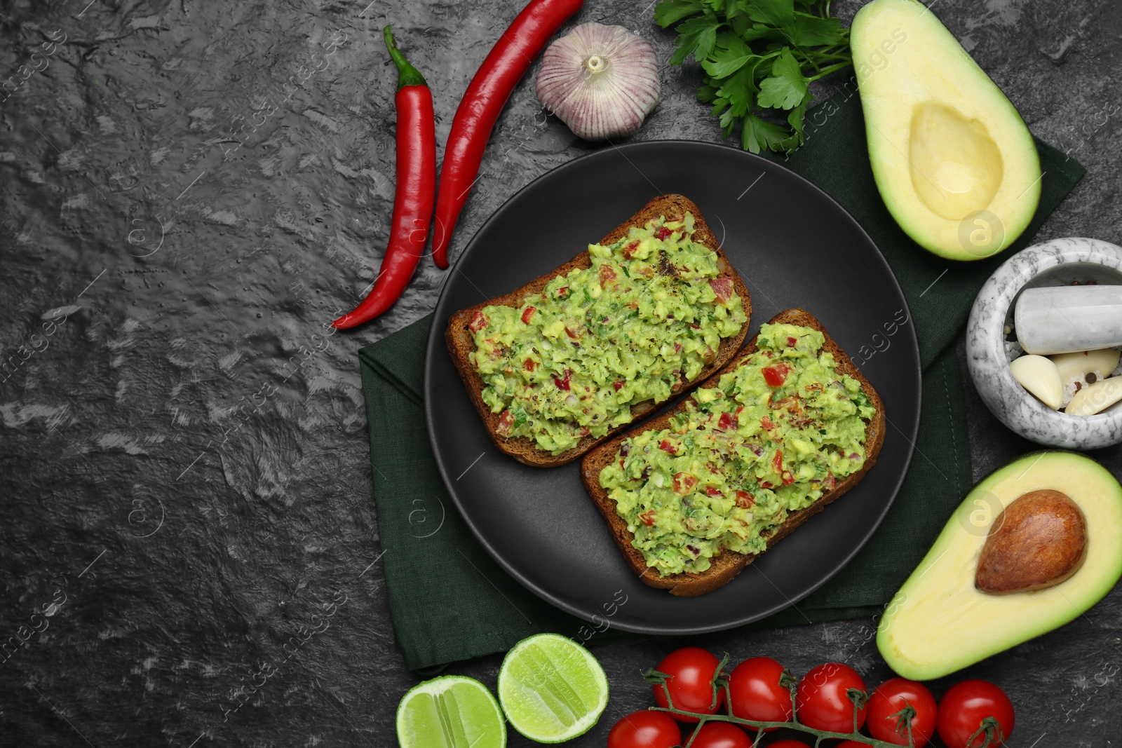 Photo of Slices of bread with tasty guacamole and ingredients on black textured table, flat lay. Space for text