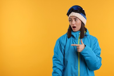 Winter sports. Surprised woman with snowboard goggles pointing at something on orange background, space for text