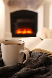 Cup of hot tea and book on knitted plaid near fireplace at home. Cozy atmosphere