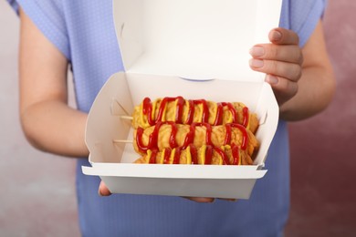 Photo of Woman holding box of delicious corn dogs with mustard and ketchup on pink background, closeup