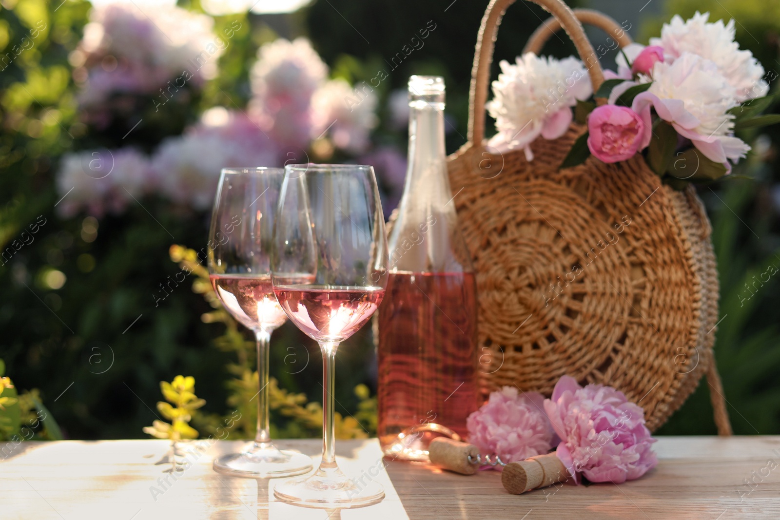 Photo of Bottle and glasses of rose wine near beautiful peonies on wooden table in garden