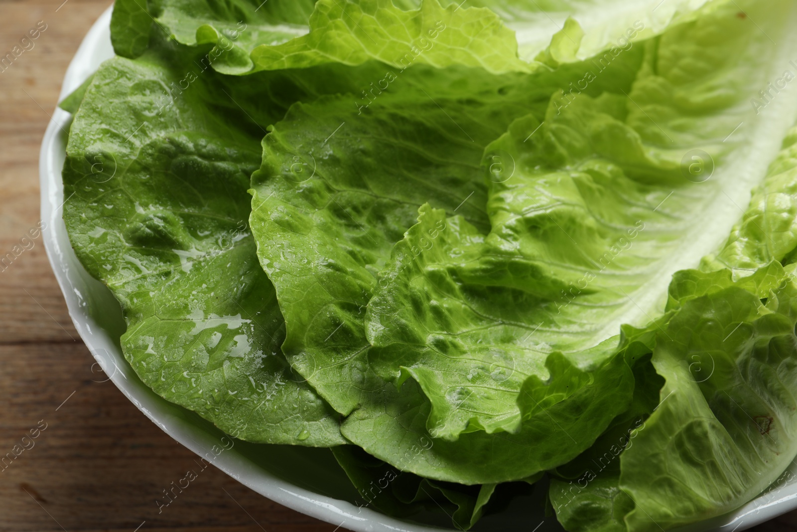 Photo of Bowl with fresh leaves of green romaine lettuce on wooden table, closeup