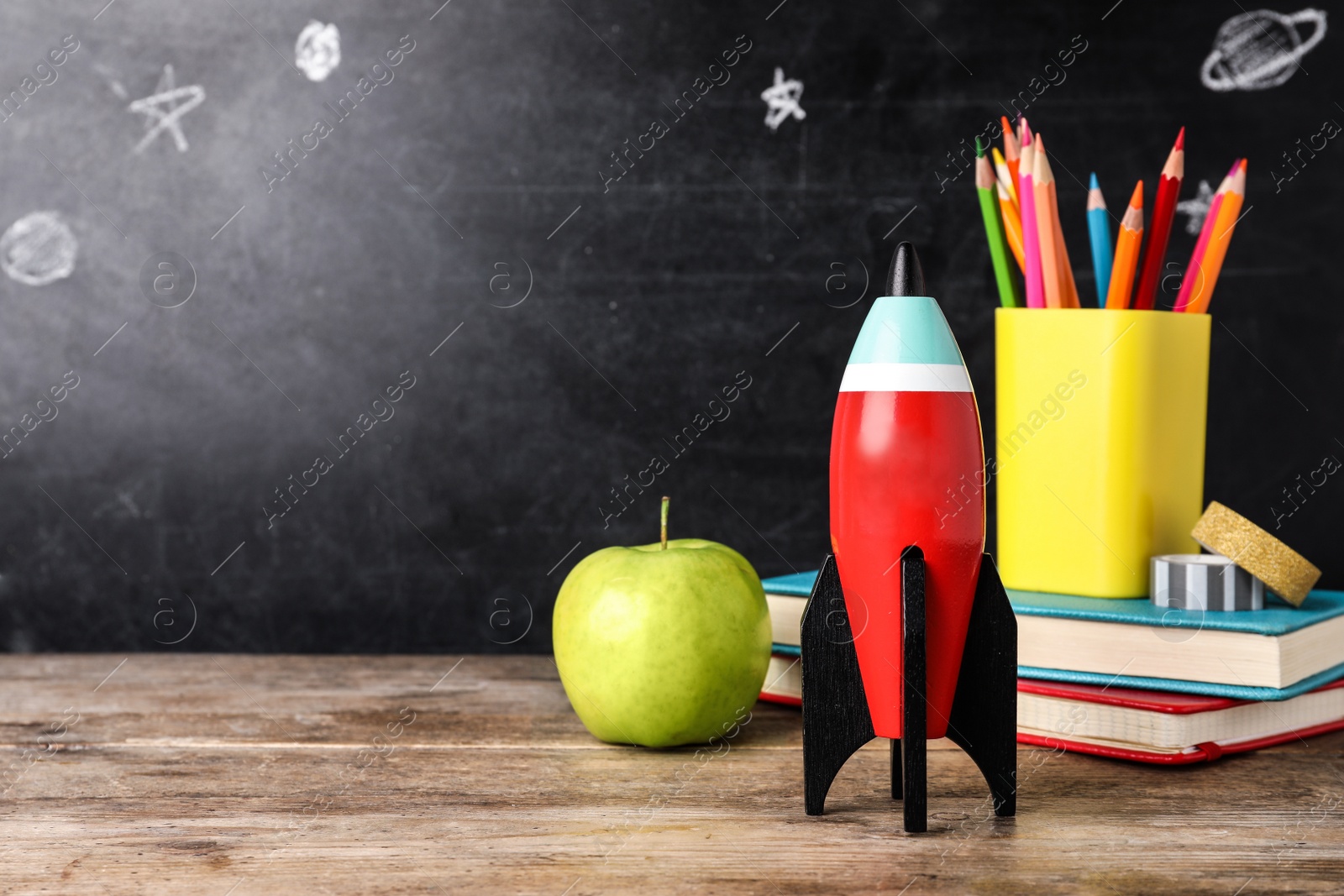 Photo of Bright toy rocket and school supplies on wooden desk. Space for text