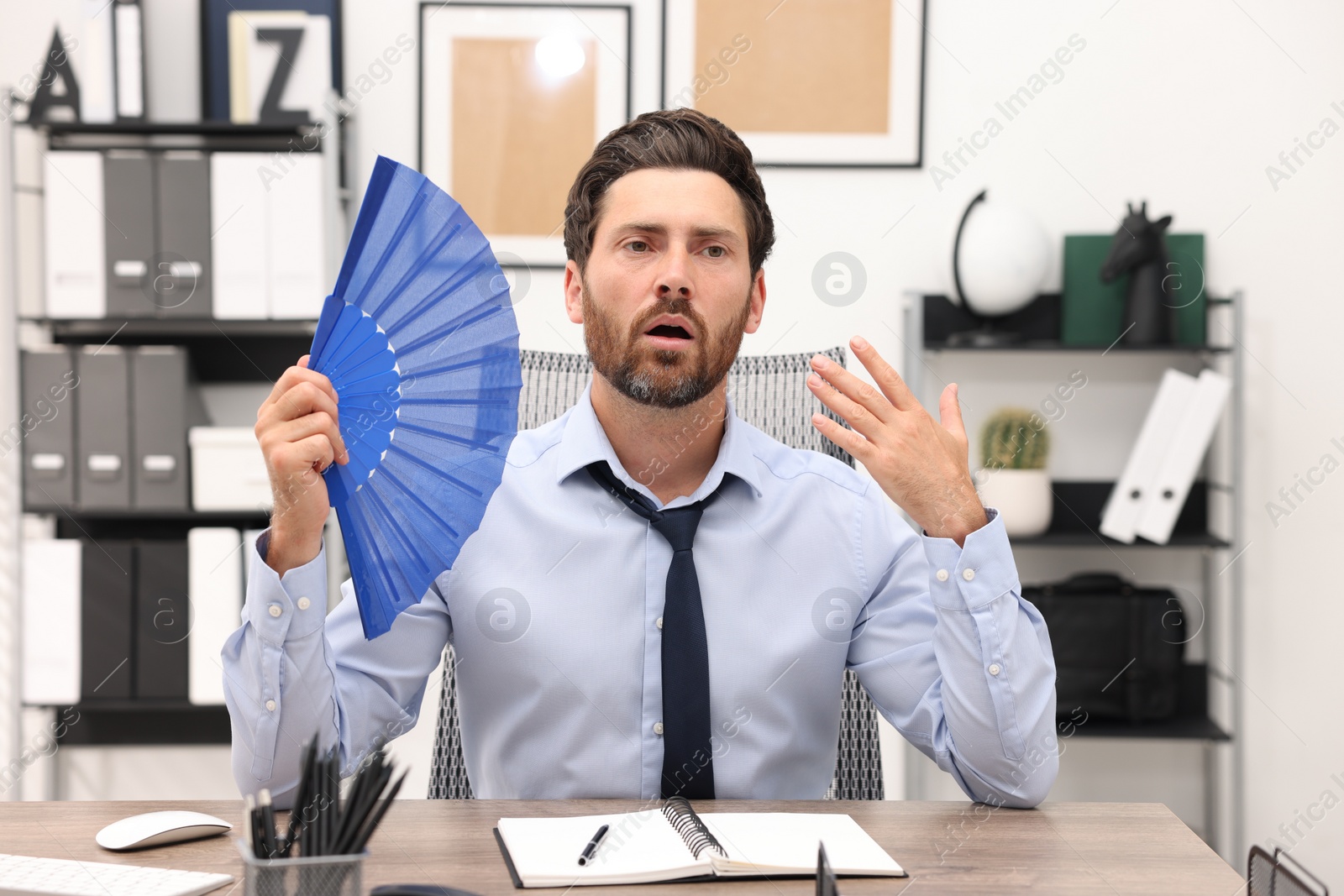 Photo of Bearded businessman waving blue hand fan to cool himself at table in office