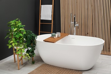 Photo of Spa day. Ceramic tub with wooden tray and green houseplants in bathroom