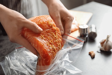 Photo of Woman putting marinated salmon fillet into plastic bag at table