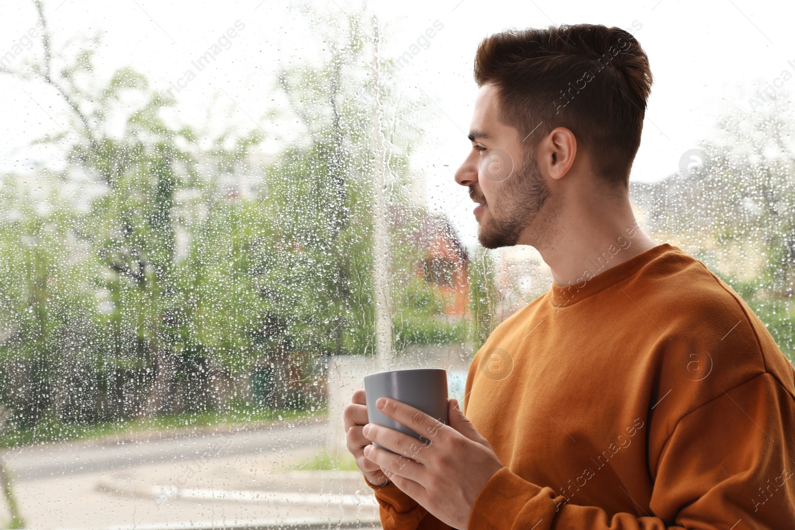 Photo of Happy handsome man with cup of coffee near window on rainy day