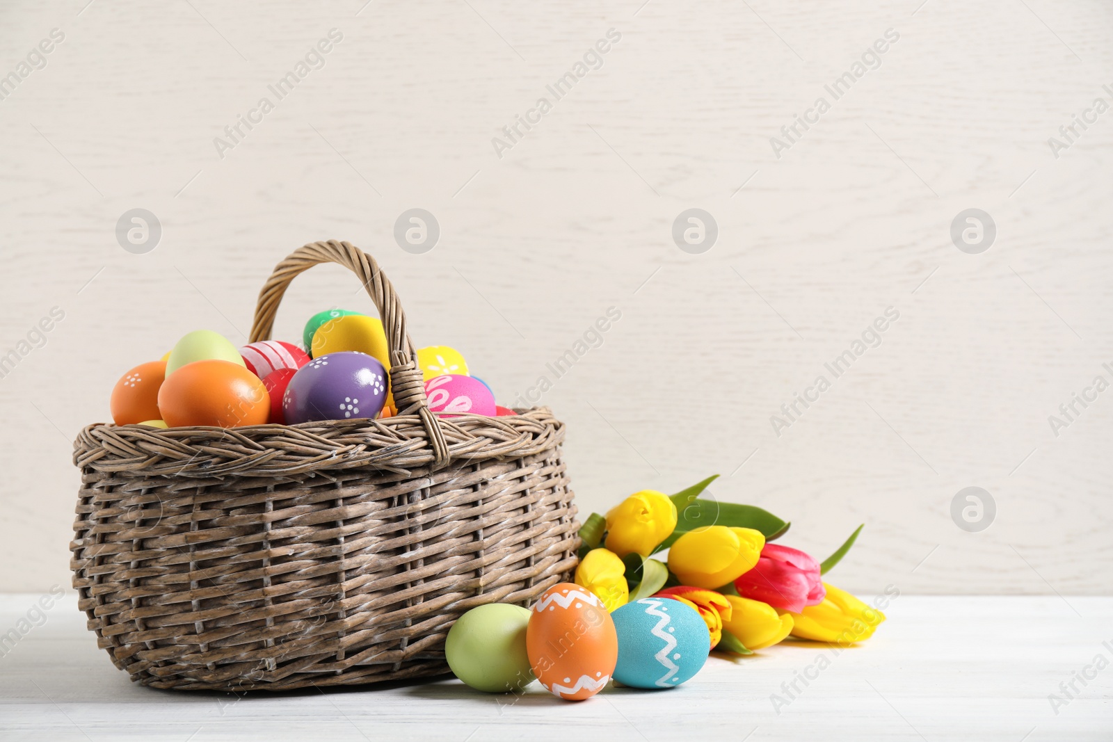 Photo of Colorful Easter eggs in basket and tulips on white wooden table. Space for text