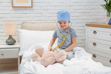 Photo of Cute child playing doctor with stuffed toy on bed in hospital ward