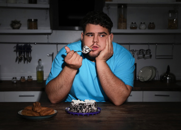 Depressed overweight man eating cake in kitchen at night