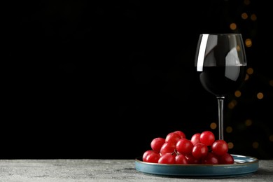 Photo of Glass of tasty red wine and fresh grapes on table against dark background with blurred lights. Space for text