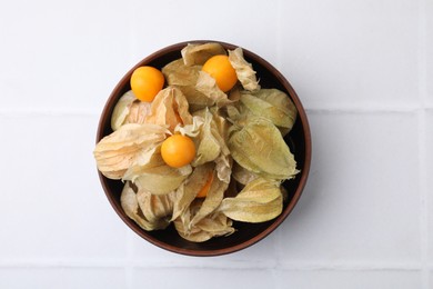 Ripe physalis fruits with calyxes in bowl on white tiled table, top view