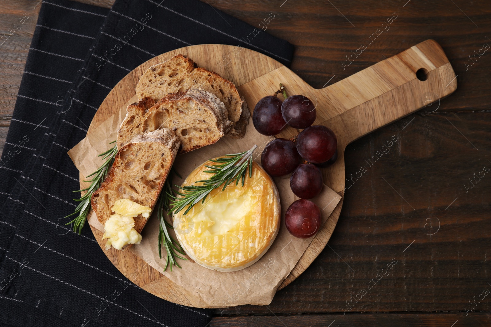 Photo of Tasty baked camembert, pieces of bread, grapes and rosemary on wooden table, top view