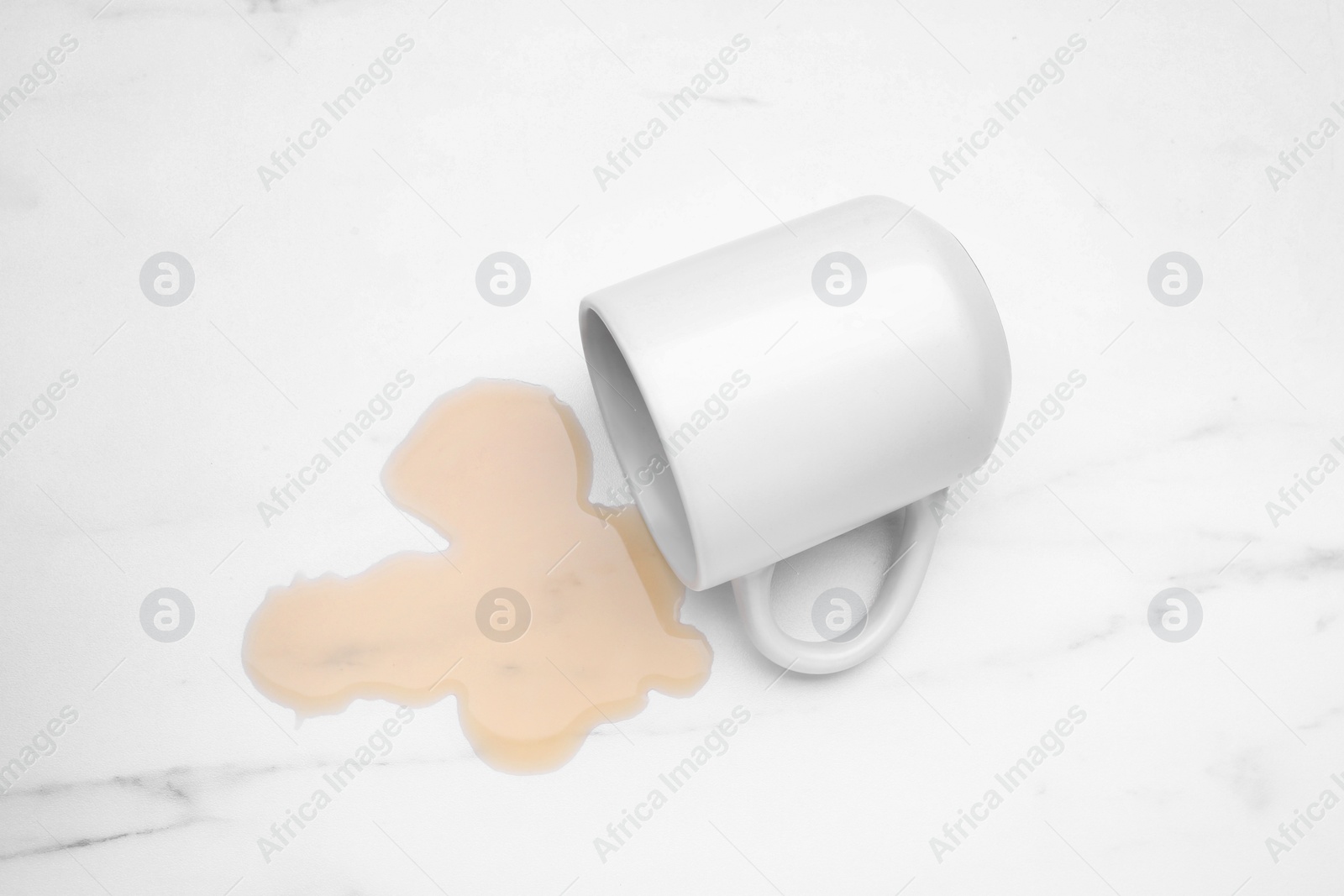 Photo of Puddle of liquid and overturned mug on white marble table, top view