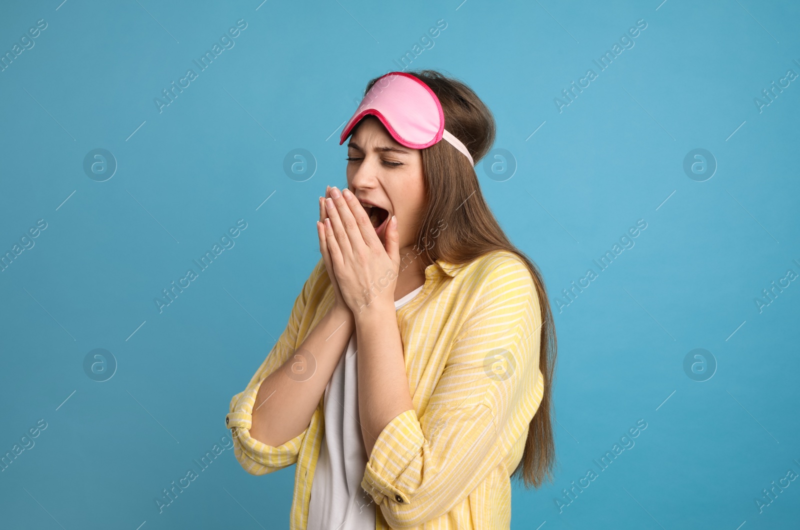 Photo of Young tired woman with sleeping mask yawning on light blue background