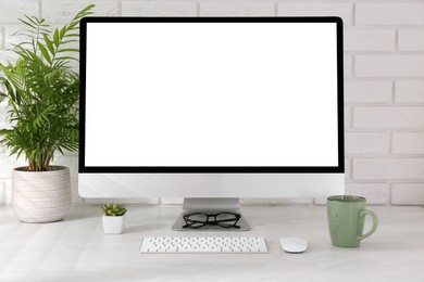 Office workplace with computer, cup, glasses, stationery and houseplant on light table near white brick wall