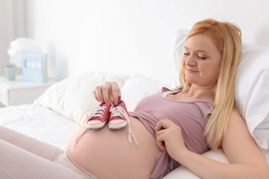 Photo of Beautiful pregnant woman holding baby booties near tummy on bed in light room