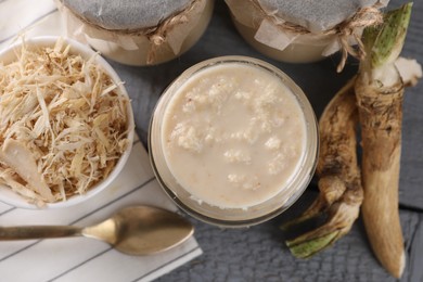 Photo of Spicy horseradish sauce in jars, roots and spoon on grey wooden table, flat lay
