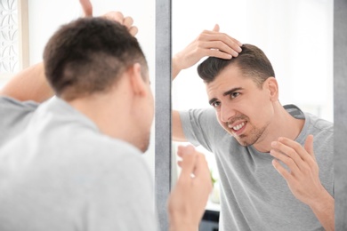 Young man with hair loss problem looking in mirror indoors