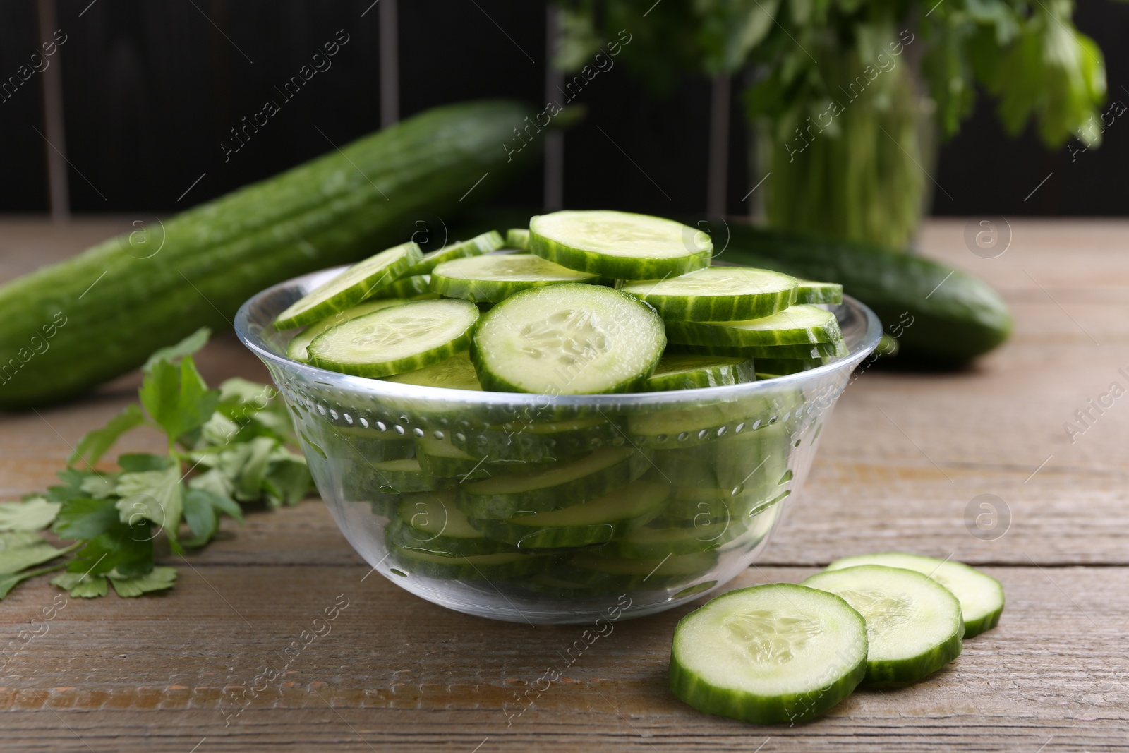 Photo of Cut cucumber in glass bowl, fresh vegetables and parsley on wooden table, closeup