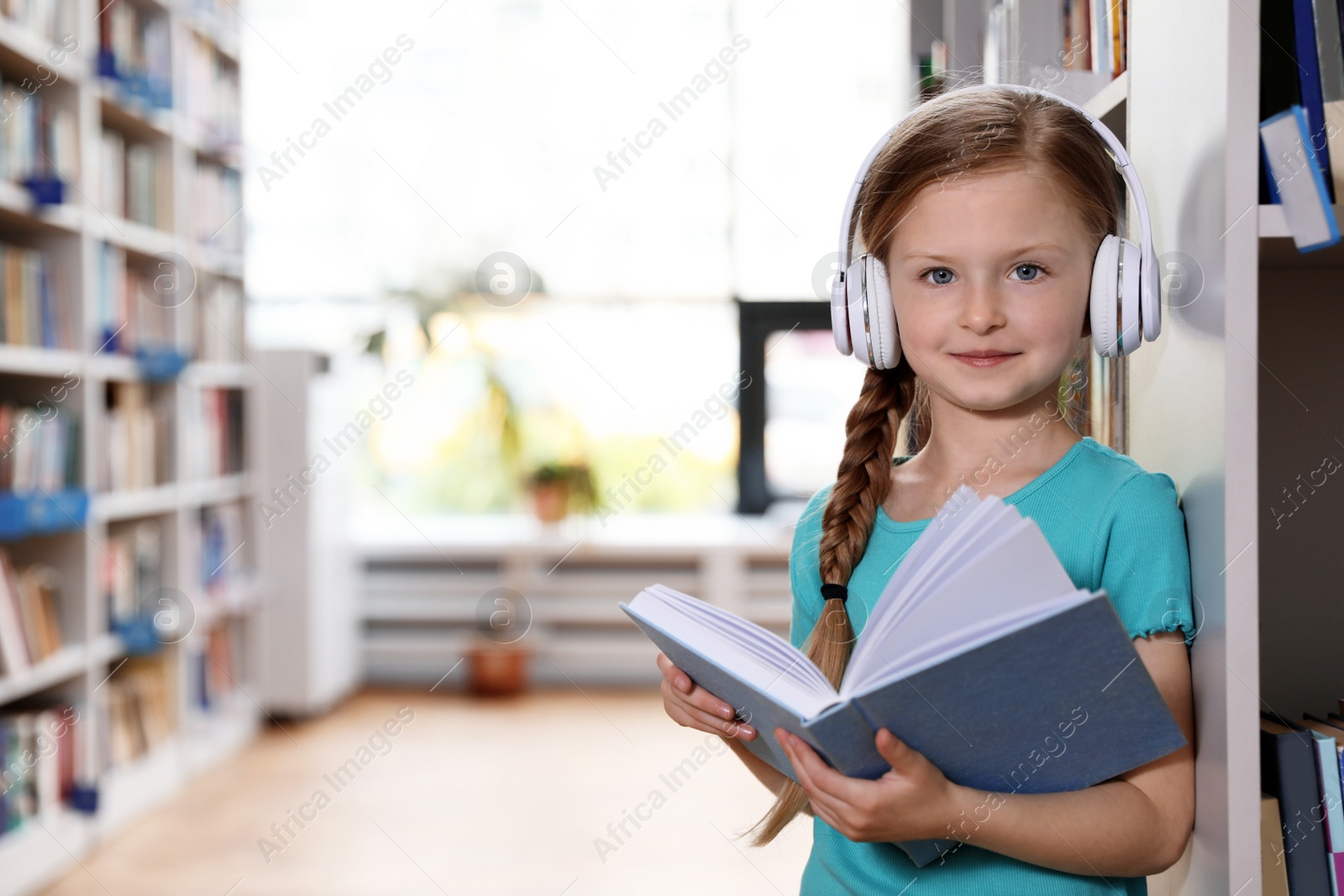 Photo of Cute little girl with headphones reading book in library