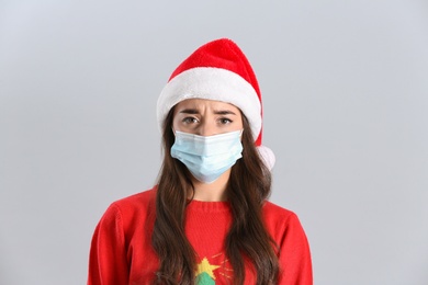 Beautiful emotional woman wearing Santa Claus hat and medical mask on light grey background