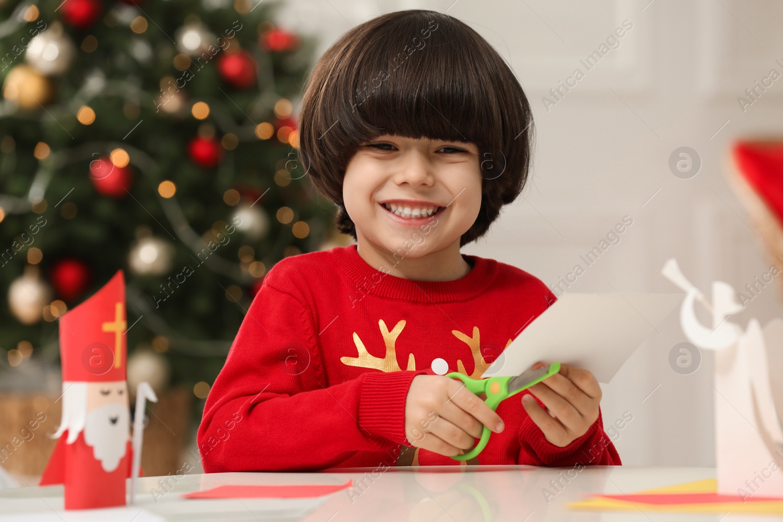 Photo of Cute little boy cutting paper at table with Saint Nicholas toy indoors