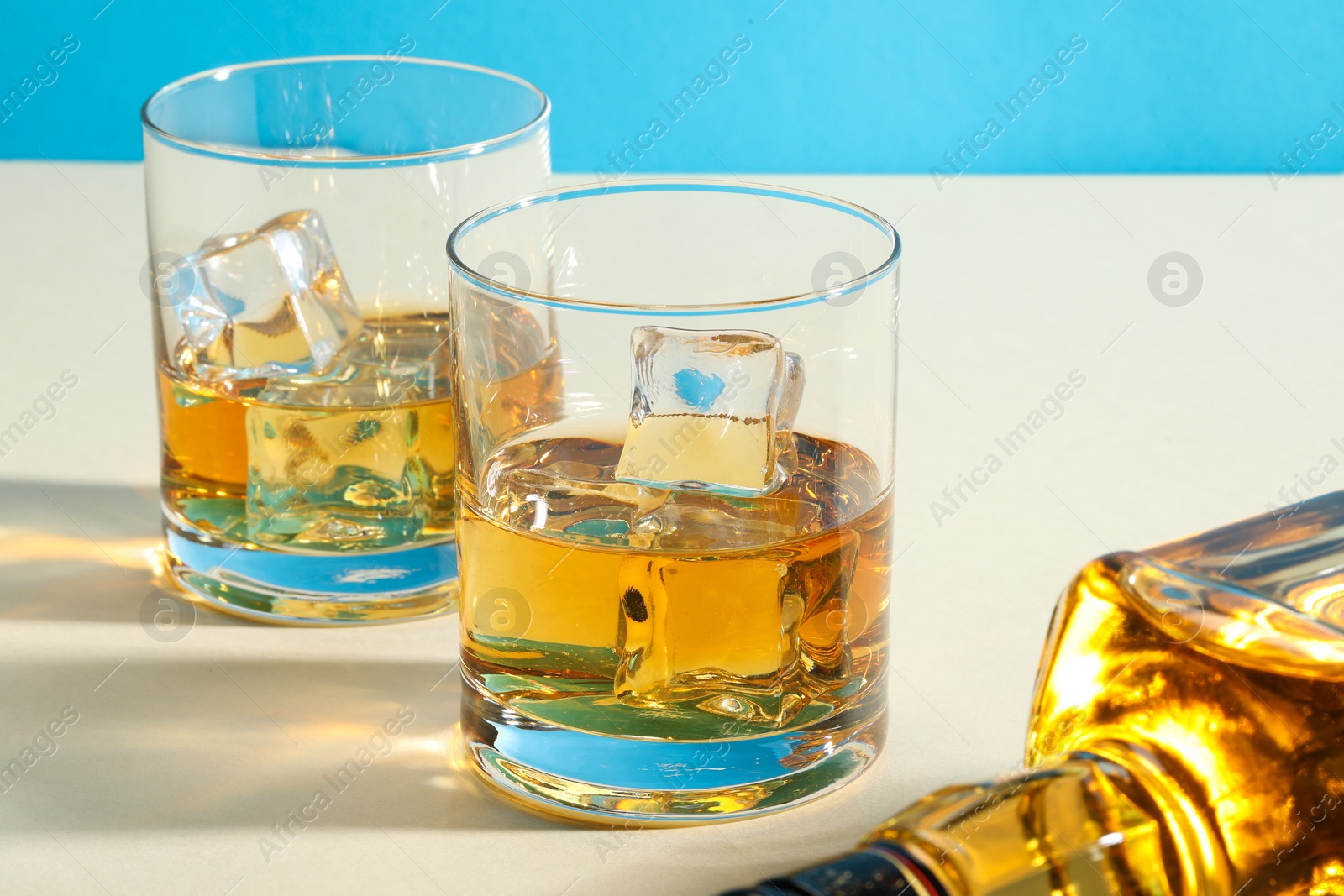 Photo of Whiskey with ice cubes in glasses and bottle on white table, closeup
