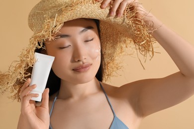 Photo of Beautiful young woman in straw hat with sunscreen on her face holding sun protection cream against beige background