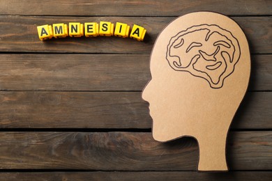Photo of Yellow cubes with word Amnesia and human head cutout with drawing of brain on wooden table, top view. Space for text
