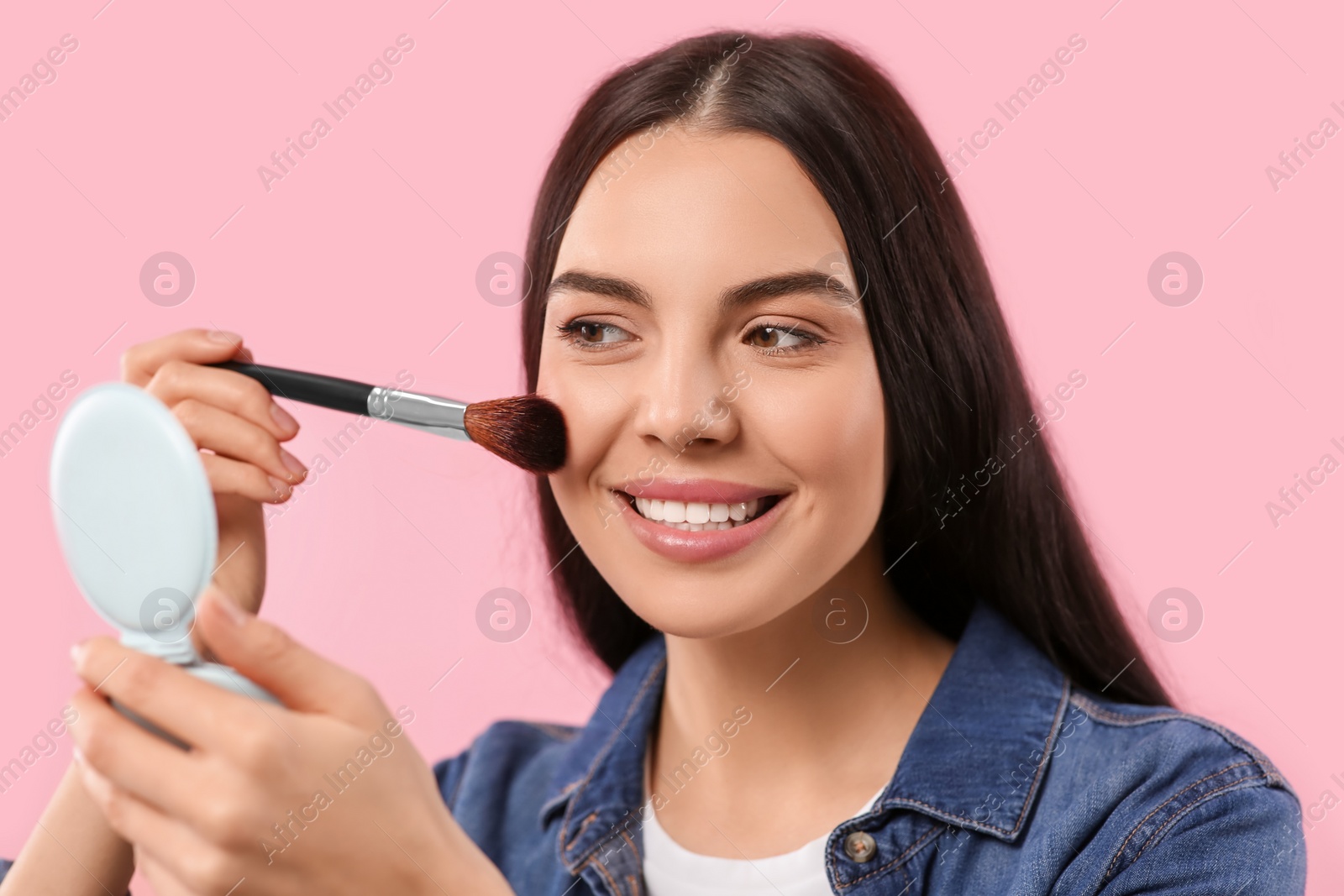 Photo of Beautiful woman with cosmetic pocket mirror applying makeup on pink background