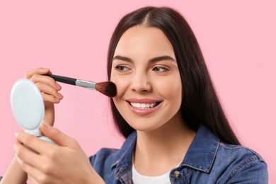 Beautiful woman with cosmetic pocket mirror applying makeup on pink background