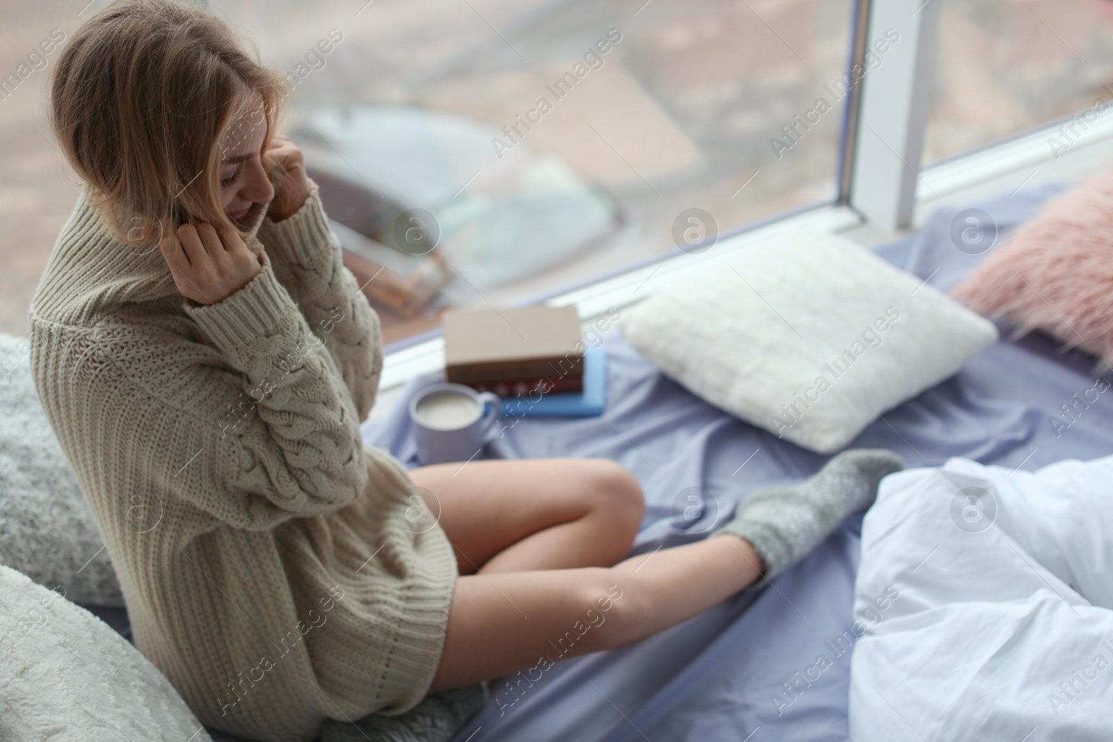 Photo of Beautiful young woman in knitted sweater sitting near window at home. Winter atmosphere