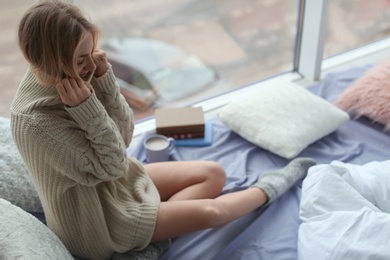 Beautiful young woman in knitted sweater sitting near window at home. Winter atmosphere