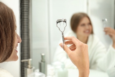 Young woman with eyelash curler near mirror in bathroom, closeup