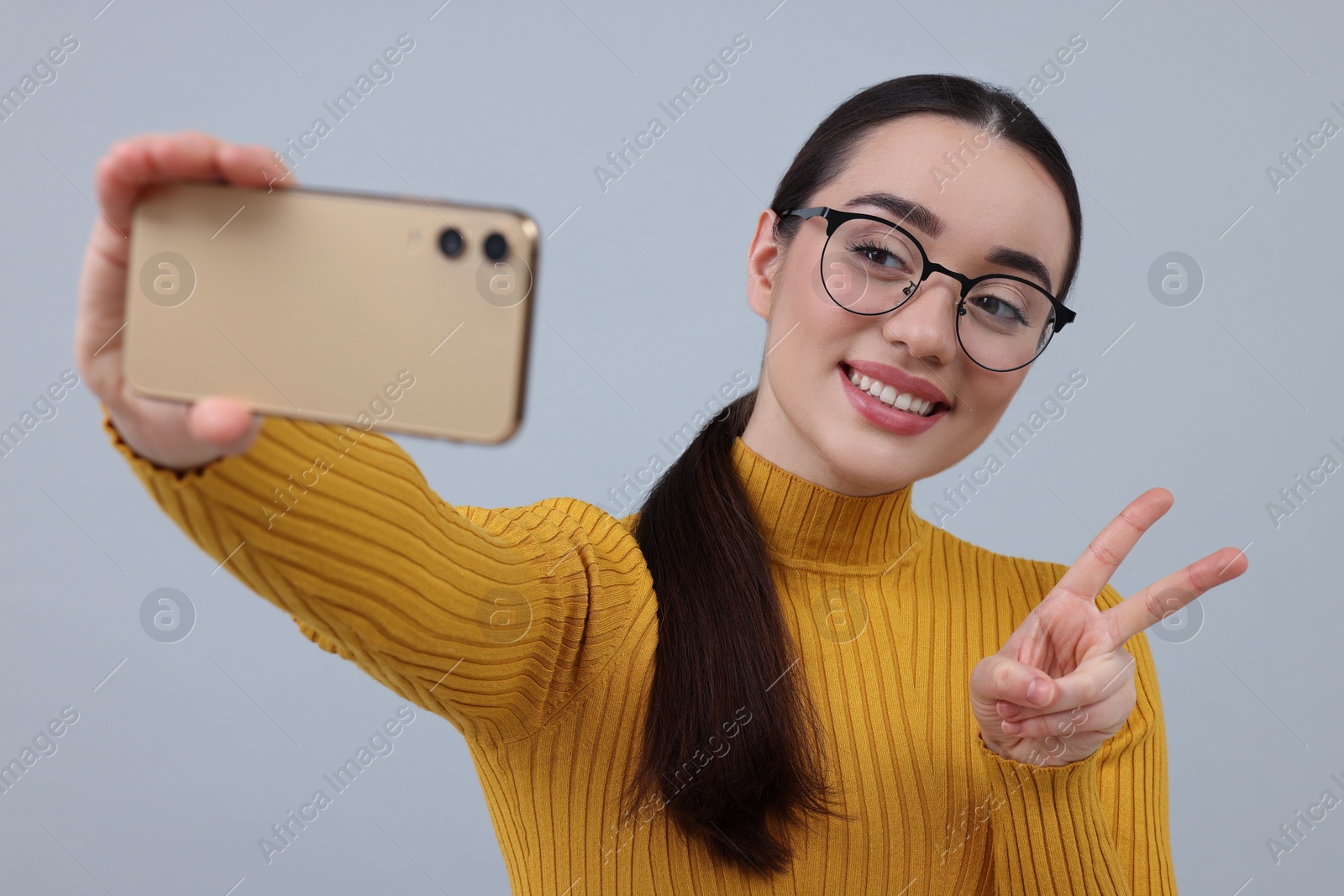 Photo of Smiling young woman taking selfie with smartphone and showing peace sign on grey background