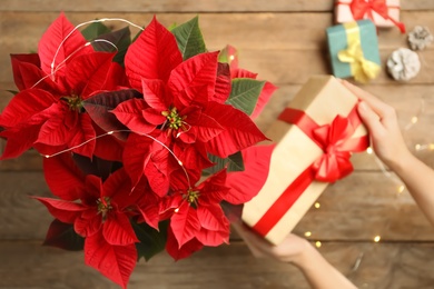 Woman with gift near poinsettia (traditional Christmas flower) at wooden table, top view