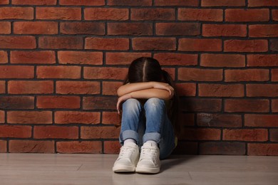 Child abuse. Upset little girl sitting on floor near brick wall indoors