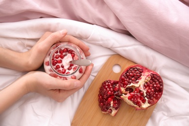 Young woman eating tasty yogurt with pomegranate seeds in bed