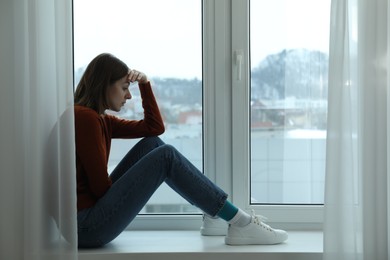 Photo of Sad young woman sitting on windowsill near window at home, space for text