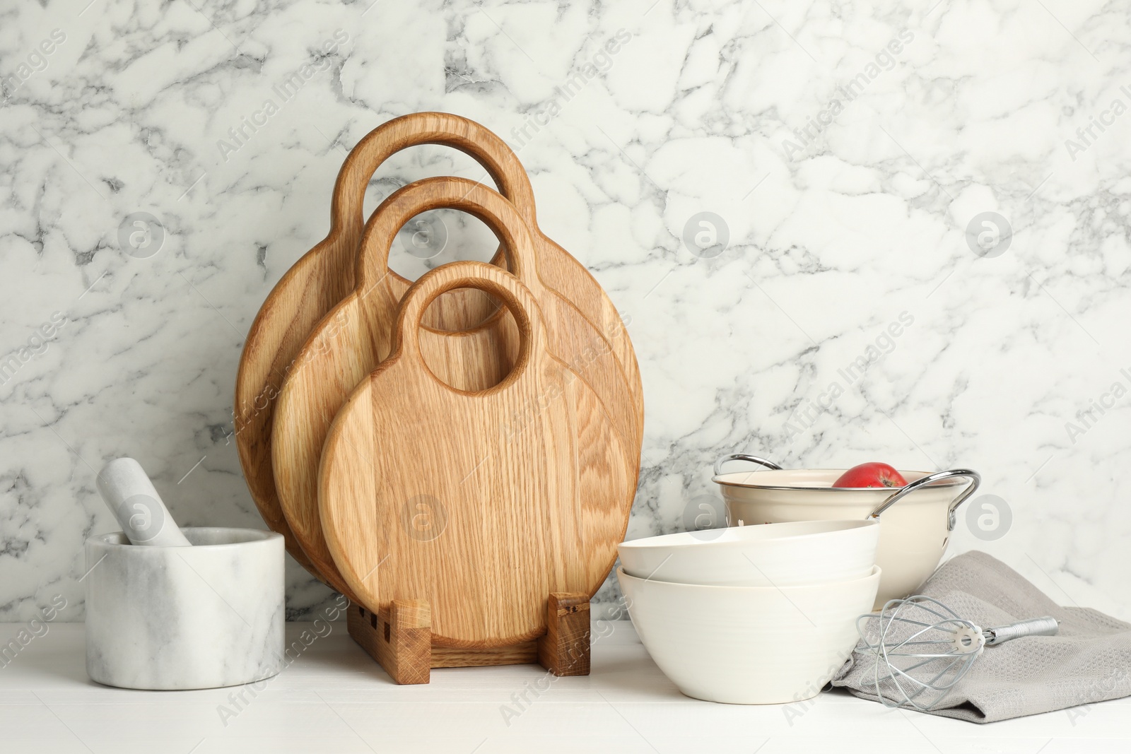 Photo of Wooden cutting boards, dishware and kitchen utensils on white table near marble wall